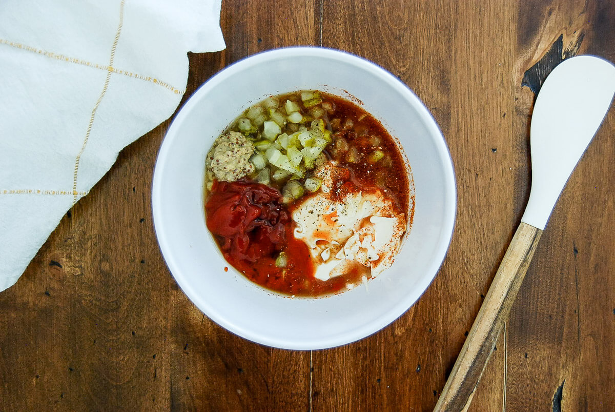 A white bowl with ketchup, chopped pickles, mayo, seasoning, and mustard sits on a wooden table next to a white cloth and a wooden spatula.