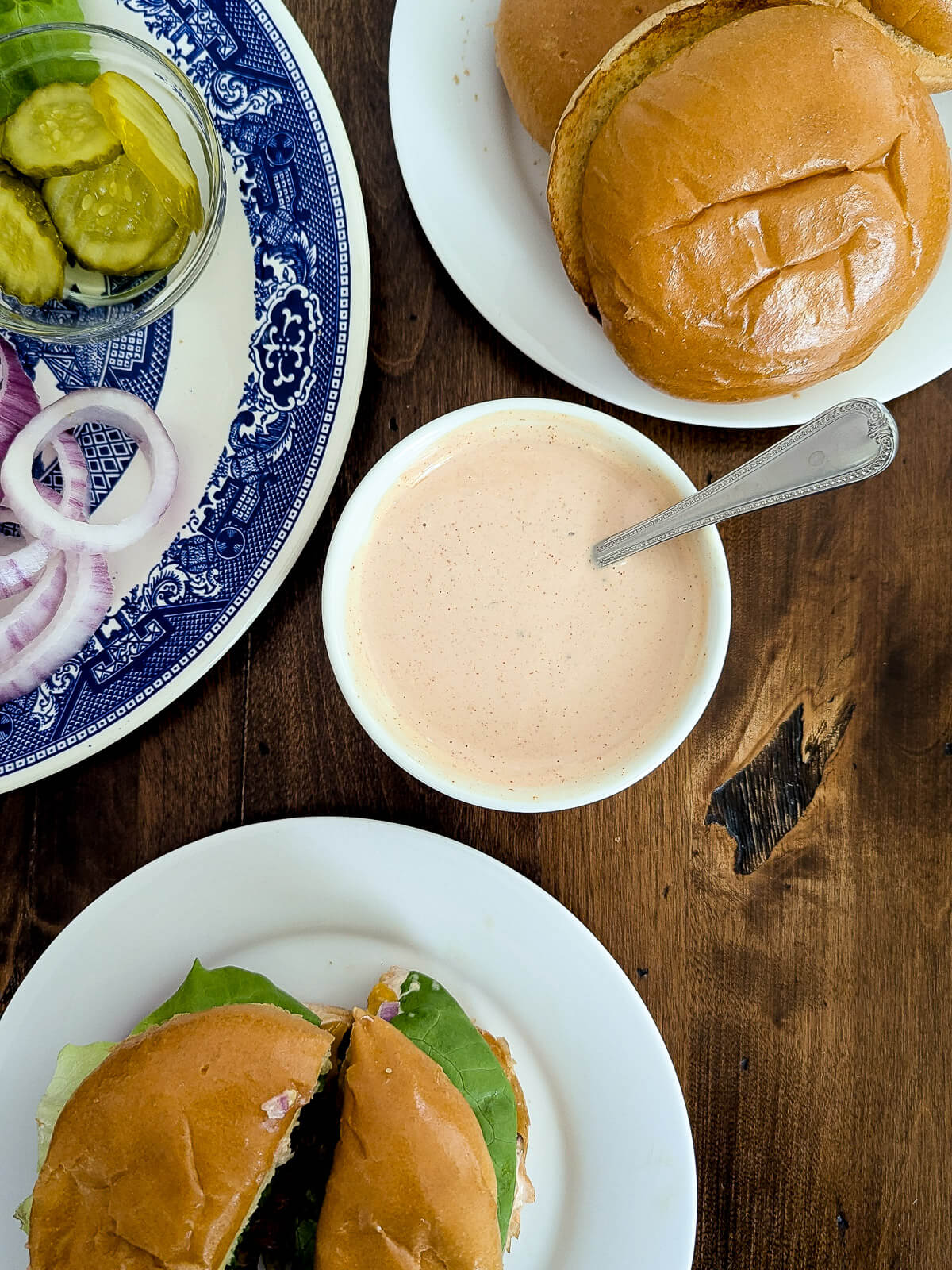 A bowl of creamy burger sauce with a spoon is surrounded by a burger bun, a sandwich with lettuce, and a plate of pickles and onion slices on a dark wooden table.