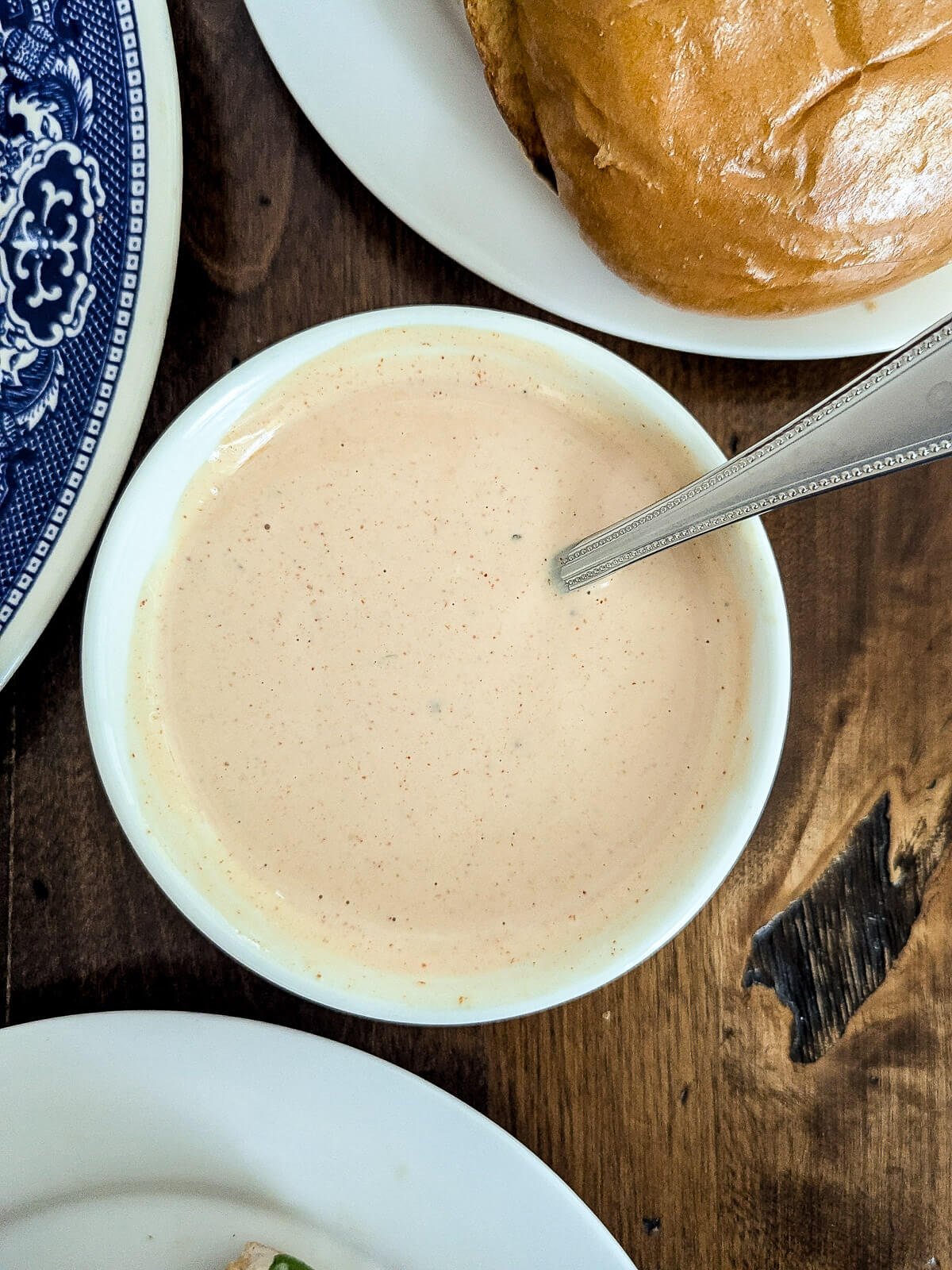 A small white bowl filled with creamy, burger sauce with specks, a spoon inside. The bowl is on a wooden table near a burger bun and a blue patterned plate.