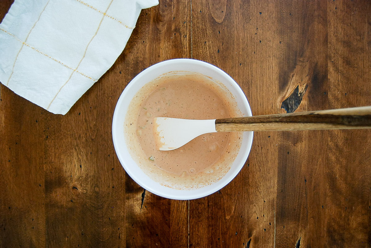 A white bowl with burger sauce being mixed by a white spatula with a wooden handle, sitting on a wooden table next to a folded white cloth.