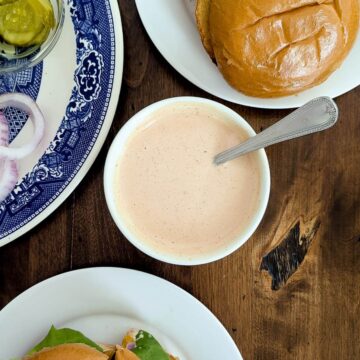 A bowl of creamy burger sauce with a spoon, surrounded by a sandwich bun, sliced onions, pickles on a blue-patterned plate, and a sandwich on a white plate, all on a wooden table.
