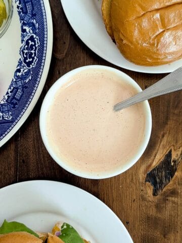 A bowl of creamy burger sauce with a spoon, surrounded by a sandwich bun, sliced onions, pickles on a blue-patterned plate, and a sandwich on a white plate, all on a wooden table.