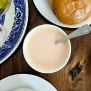 A bowl of creamy burger sauce with a spoon sits between plates with burgers, pickles, and onions on a wooden table.