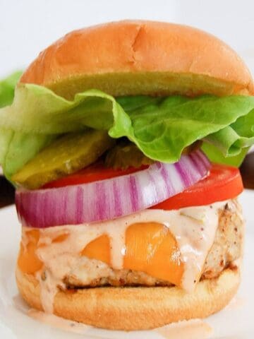 A chicken burger with cheddar cheese, lettuce, tomato, pickles, red onion, and burger sauce on a soft bun, served on a white plate.