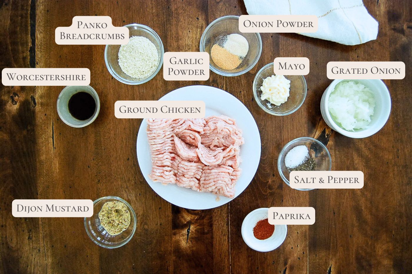 Ingredients for chicken patties on a table: ground chicken, panko breadcrumbs, garlic powder, onion powder, mayo, grated onion, salt, pepper, paprika, Dijon mustard, and Worcestershire sauce in bowls.