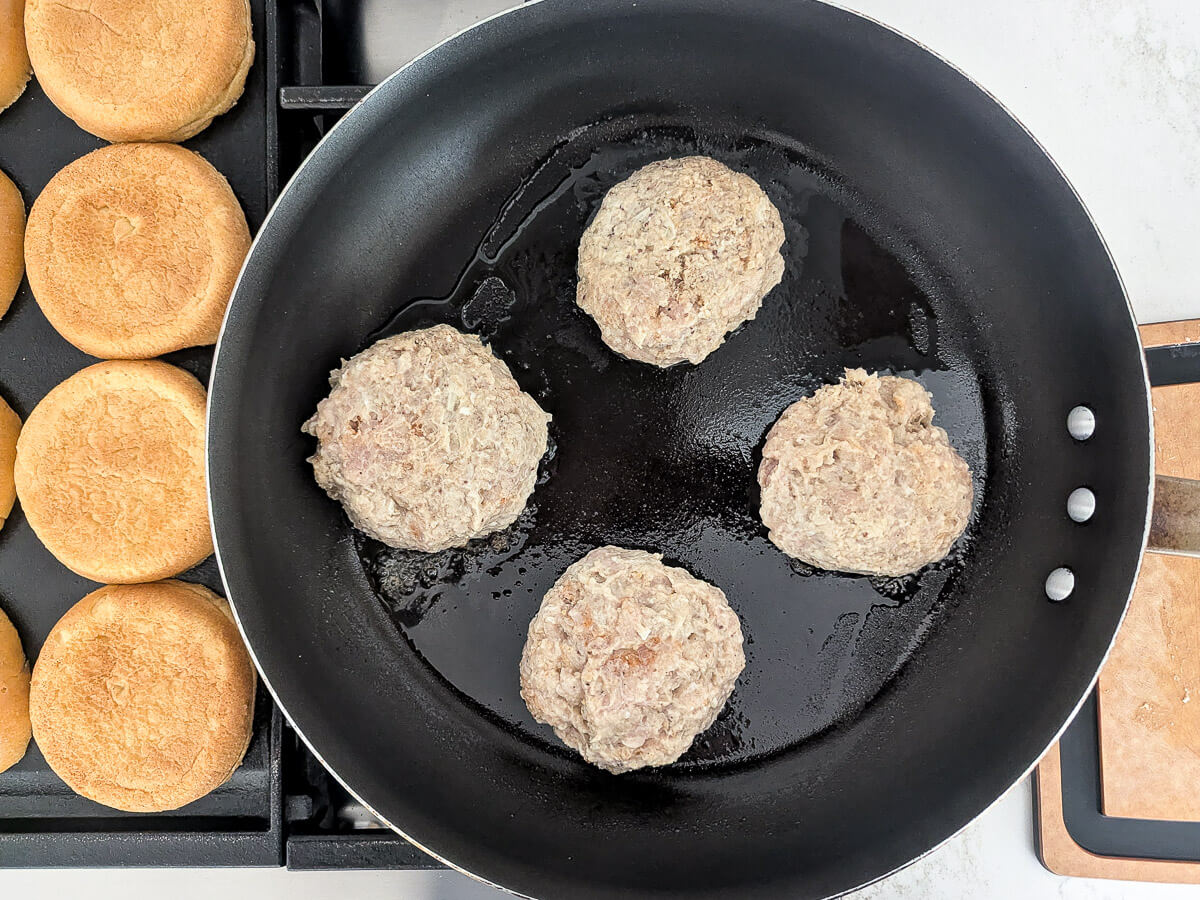 Four raw chicken burger patties are cooking in a black skillet on a stove. To the left, several burger buns are toasting on the griddle. A spatula rests nearby.