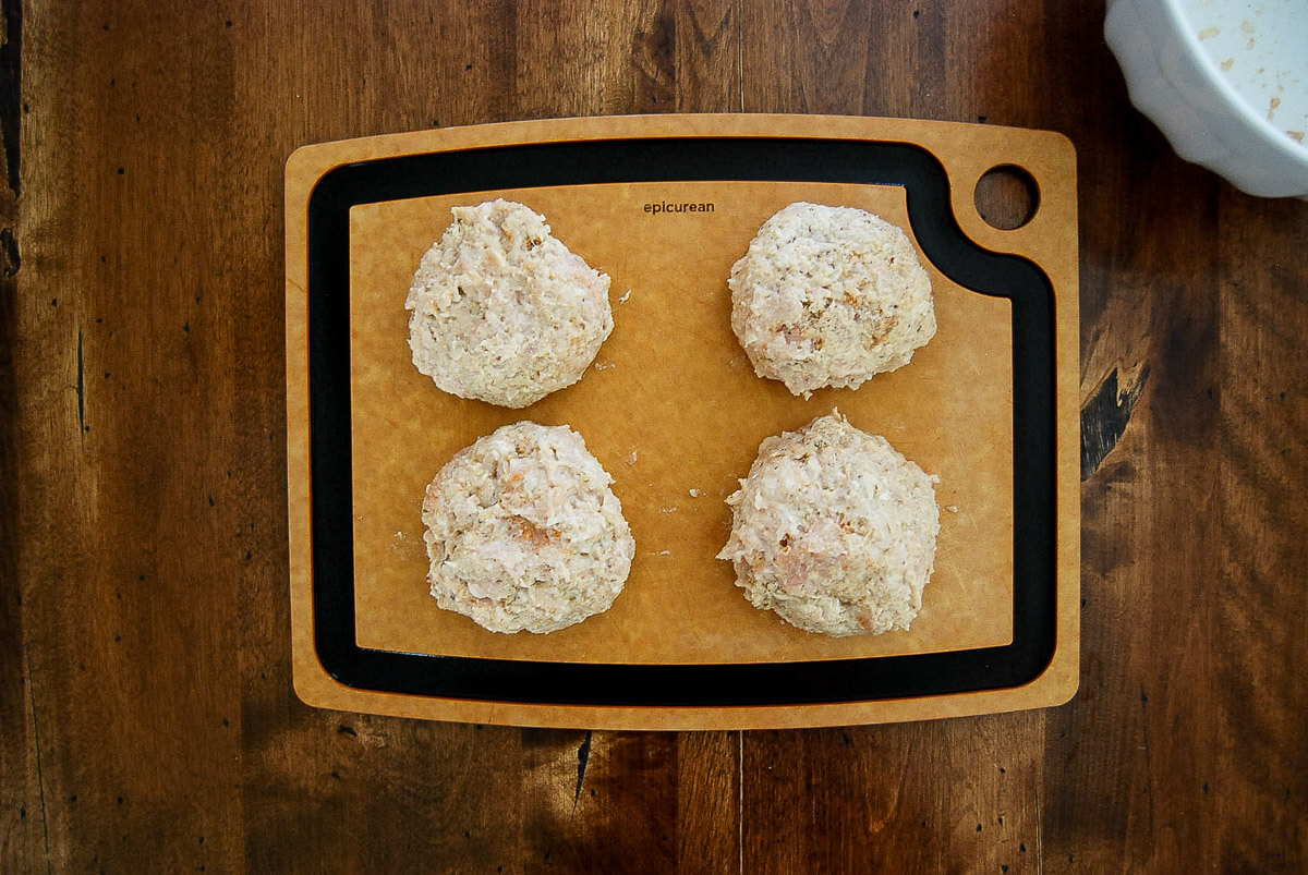Four formed chicken burger patties are sitting on a cutting board on a wooden table.