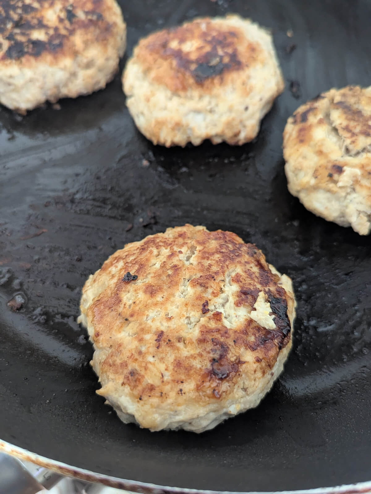 Four browned chicken burgers are being cooked on a black pan.