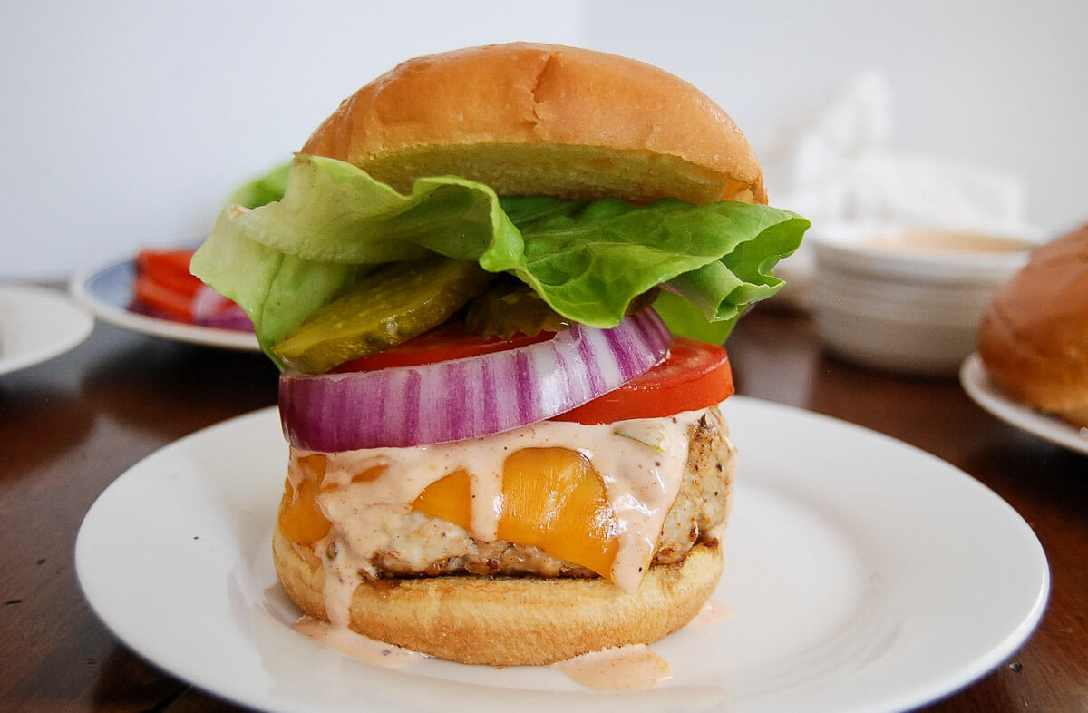 A chicken burger with lettuce, tomato, onion, pickles, cheddar cheese, and sauce on a bun, served on a white plate.