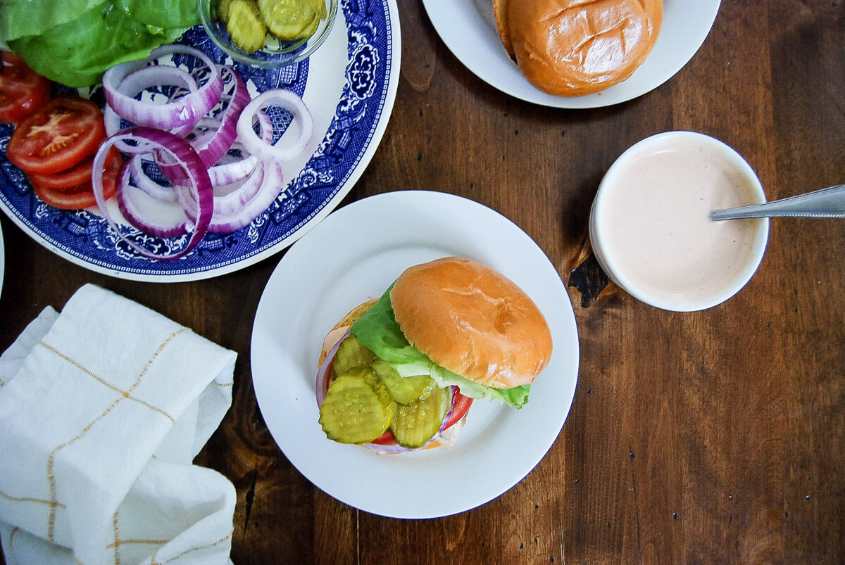 Open chicken burger with lettuce, pickles, tomato, and onion on a plate, beside a bowl of sauce and a plate of sliced veggies, all on a wooden table.