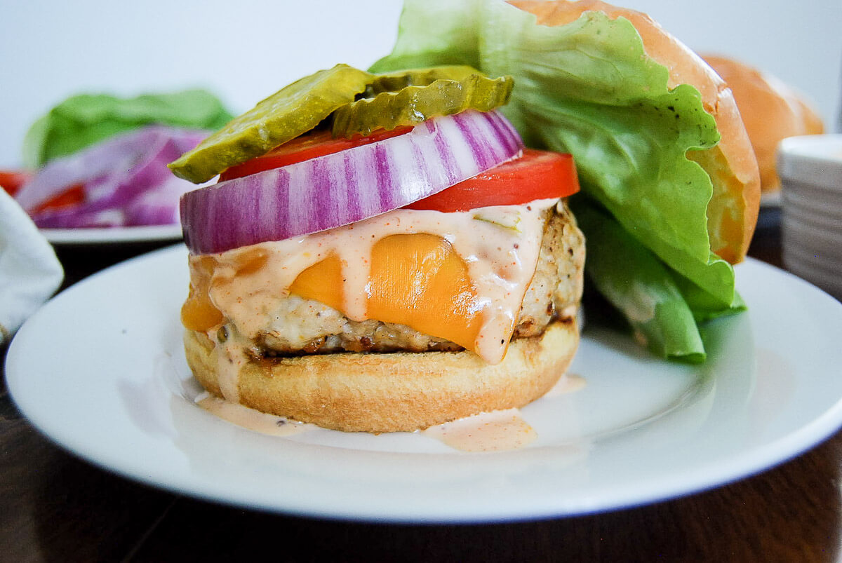 A close-up of a chicken burger with cheddar cheese, lettuce, tomato, red onion, pickles, and sauce on a white plate.