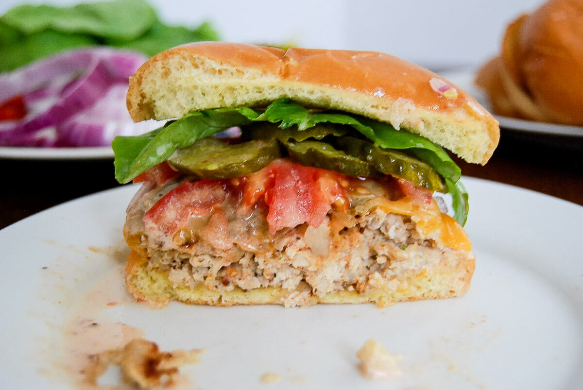 A close-up of a half-eaten chicken burger with lettuce, tomato, pickles, and burger sauce on a plate. Sliced onions and other toppings are blurred in the background.