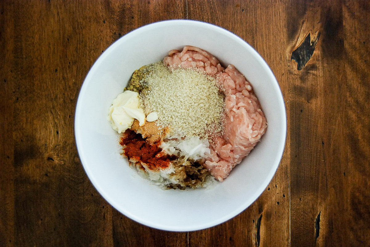 A white bowl on a wooden table filled with ground chicken, panko breadcrumbs, mayonnaise, chopped onions, and various spices before mixing.