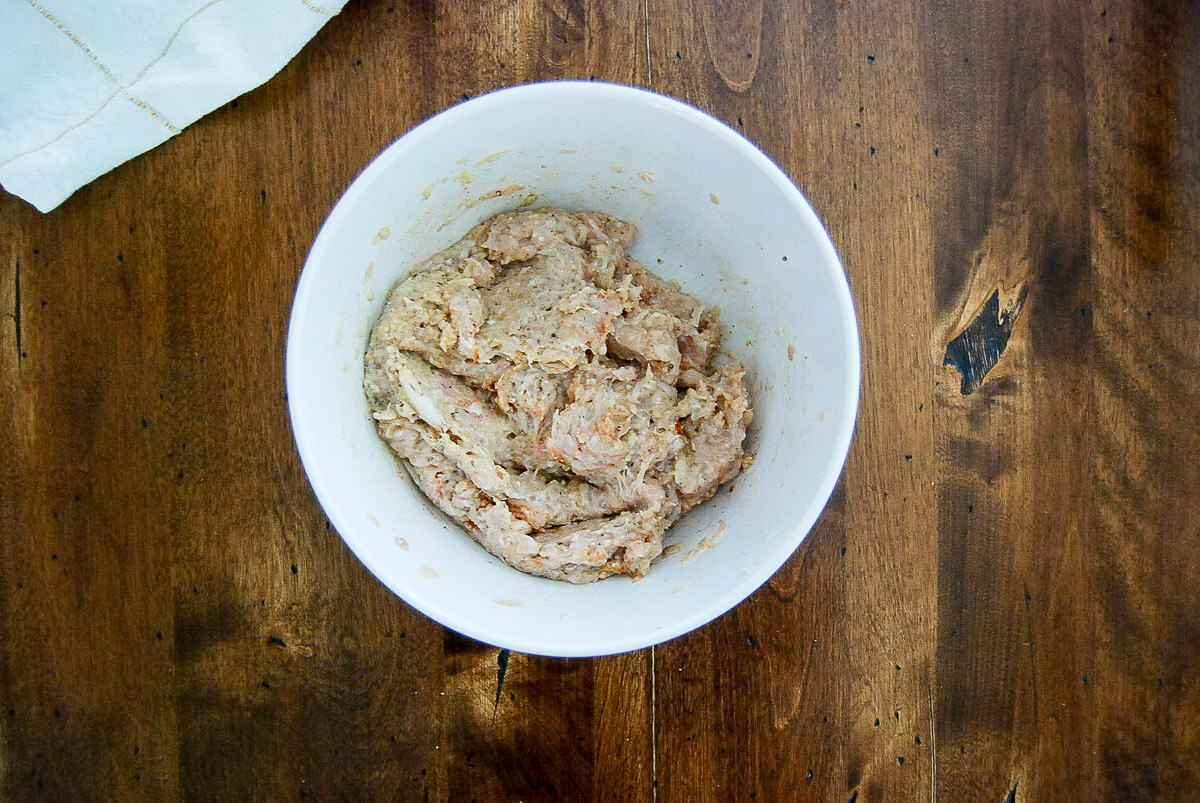 A white bowl containing a mixture of raw marinated ground chicken on a wooden surface.