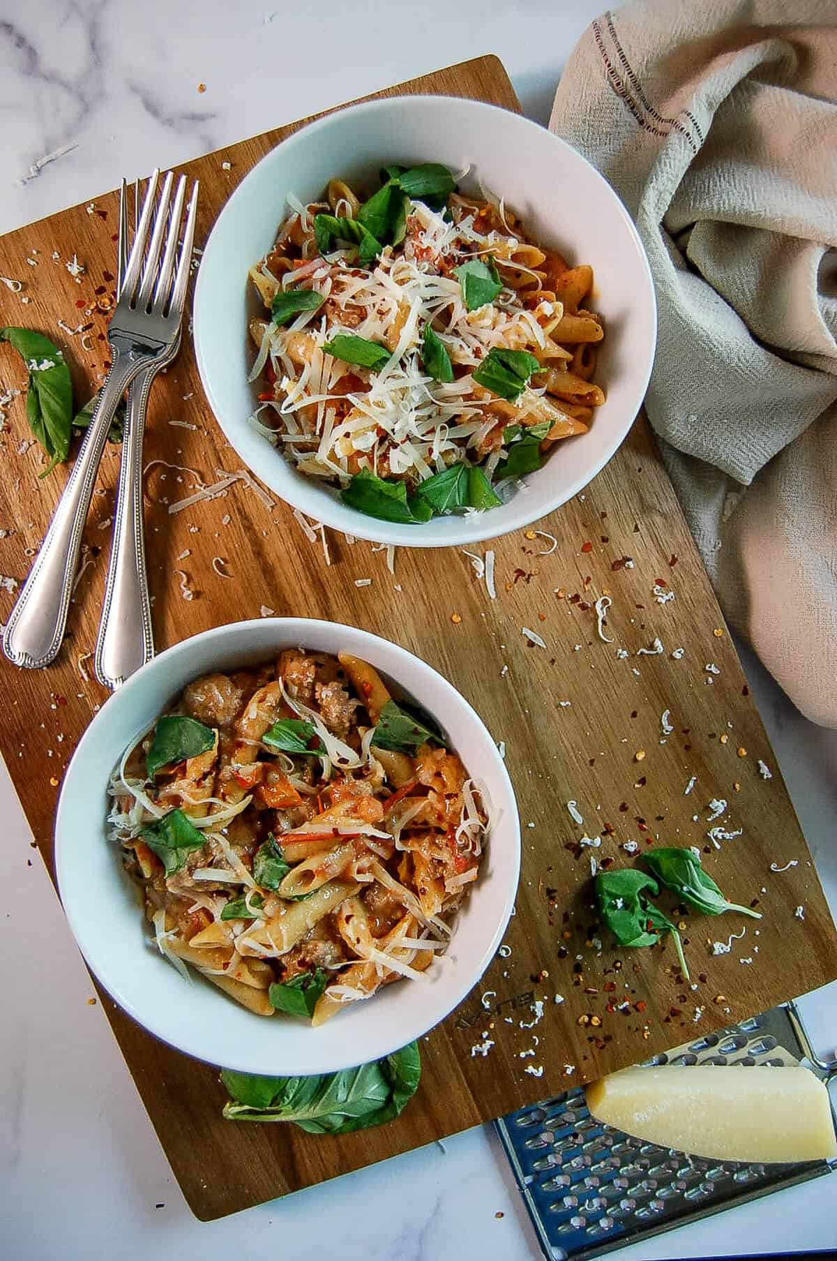 Two bowls of penne pasta with tomato sauce, Italian sausage, shredded cheese, and fresh basil on a wooden board, with forks, a grater, parmesan, and a beige napkin nearby.