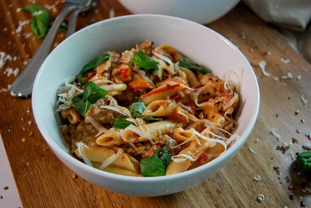 A white bowl of penne pasta with Italian sausage, tomato sauce, fresh basil, and grated Parmesan cheese sits on a wooden surface, surrounded by scattered cheese and herbs.