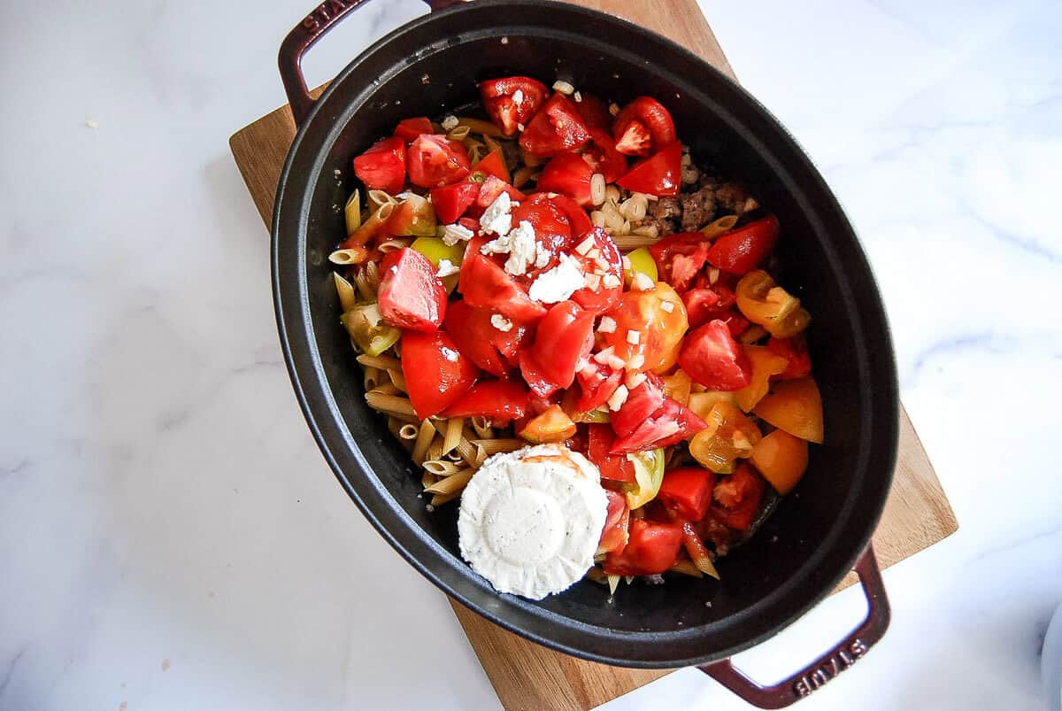 A baking dish filled with uncooked penne pasta, chopped tomatoes, bell peppers, garlic, and a round of Boursin cheese cheese, set on a wooden board on a white surface.