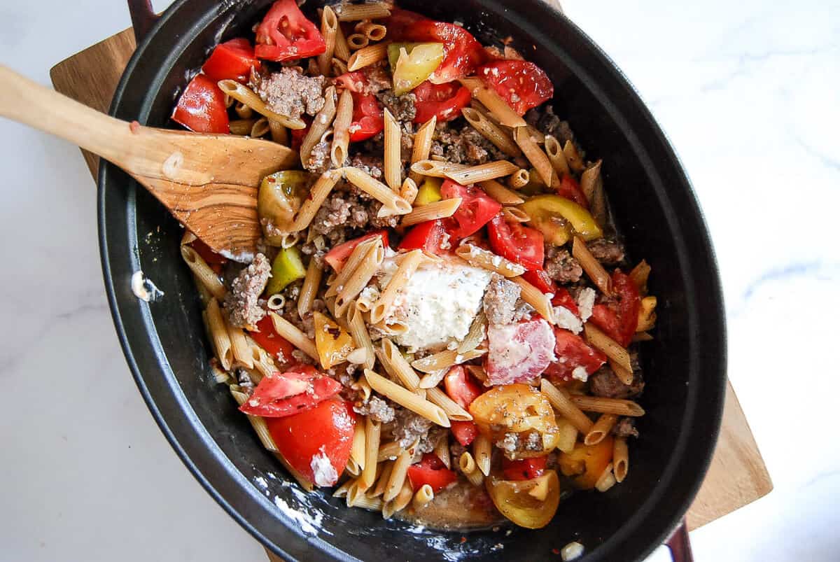 A black pot filled with penne pasta, ground meat, chopped tomatoes, bell peppers, and dollops of creamy cheese, being stirred with a wooden spoon on a light surface.