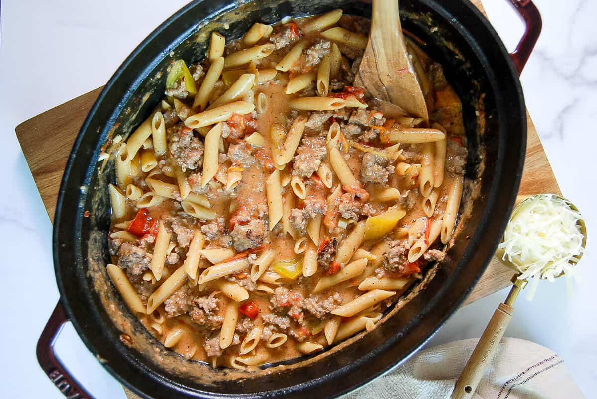 A black pot filled with penne pasta, ground meat, diced tomatoes, and sauce sits on a wooden board. A wooden spoon rests inside, and a small bowl of shredded cheese is nearby.