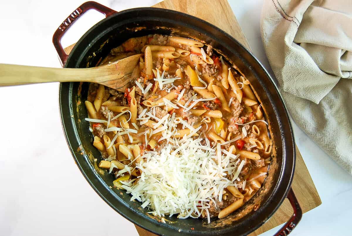A black dish filled with cooked penne pasta, meat sauce, and topped with shredded cheese. A wooden spoon rests in the dish, which sits on a wooden board next to a beige cloth napkin.