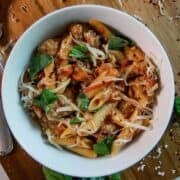 A white bowl filled with penne pasta, sausage, tomato sauce, shredded cheese, and fresh basil leaves, sits on a wooden surface with scattered herbs and cheese around it.