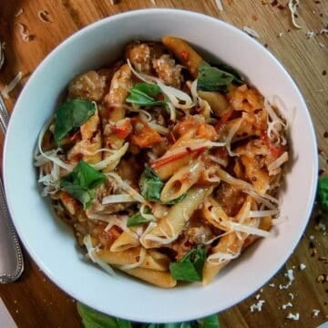 A white bowl filled with penne pasta, sausage, tomato sauce, shredded cheese, and fresh basil leaves, sits on a wooden surface with scattered herbs and cheese around it.