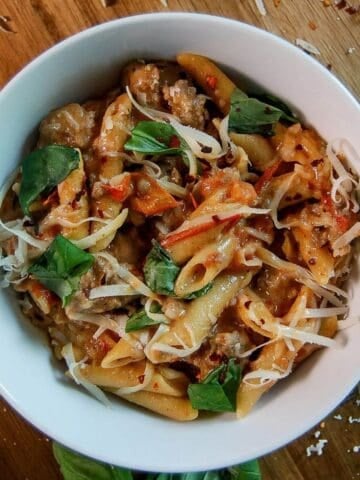 A white bowl filled with penne pasta, sausage, tomato sauce, shredded cheese, and fresh basil leaves, sits on a wooden surface with scattered herbs and cheese around it.