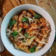 A white bowl filled with penne pasta in tomato sauce, sausage, fresh basil leaves, and shredded cheese, placed on a wooden surface with scattered cheese and basil.