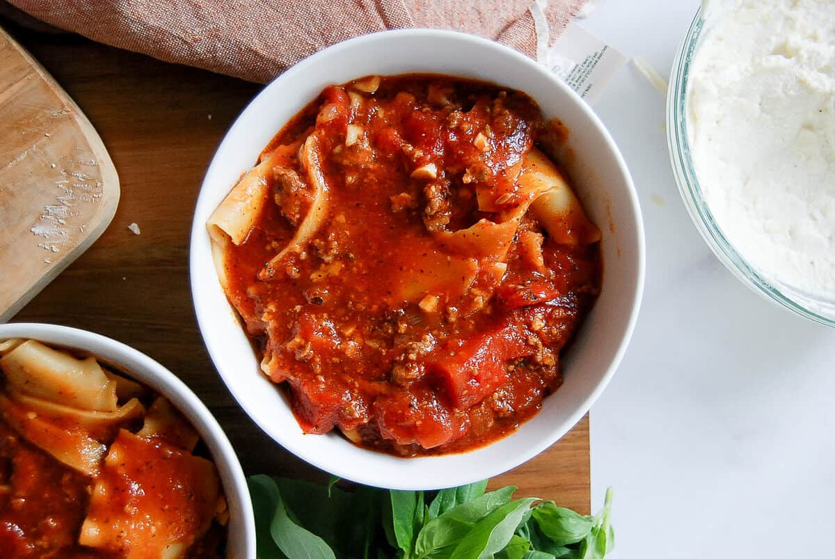 Two white bowls filled with lasagna soup topped with rich tomato sauce and beef, next to a bowl of ricotta cheese, a bunch of fresh basil, and a wooden cutting board.