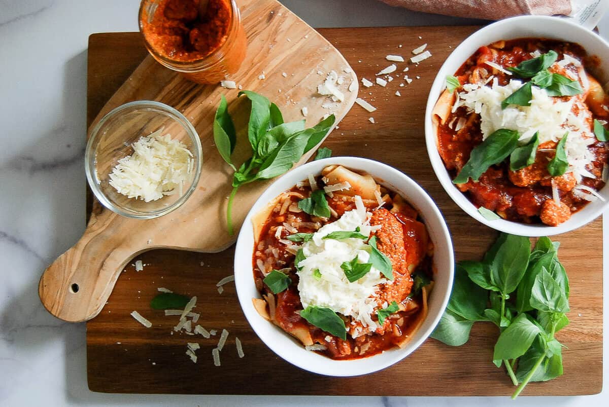 Two bowls of lasagna soup with tomato sauce, whipped ricotta, grated cheese, and fresh basil on a wooden board. A jar of red pepper sauce, a bowl of grated cheese, and basil leaves are arranged nearby.