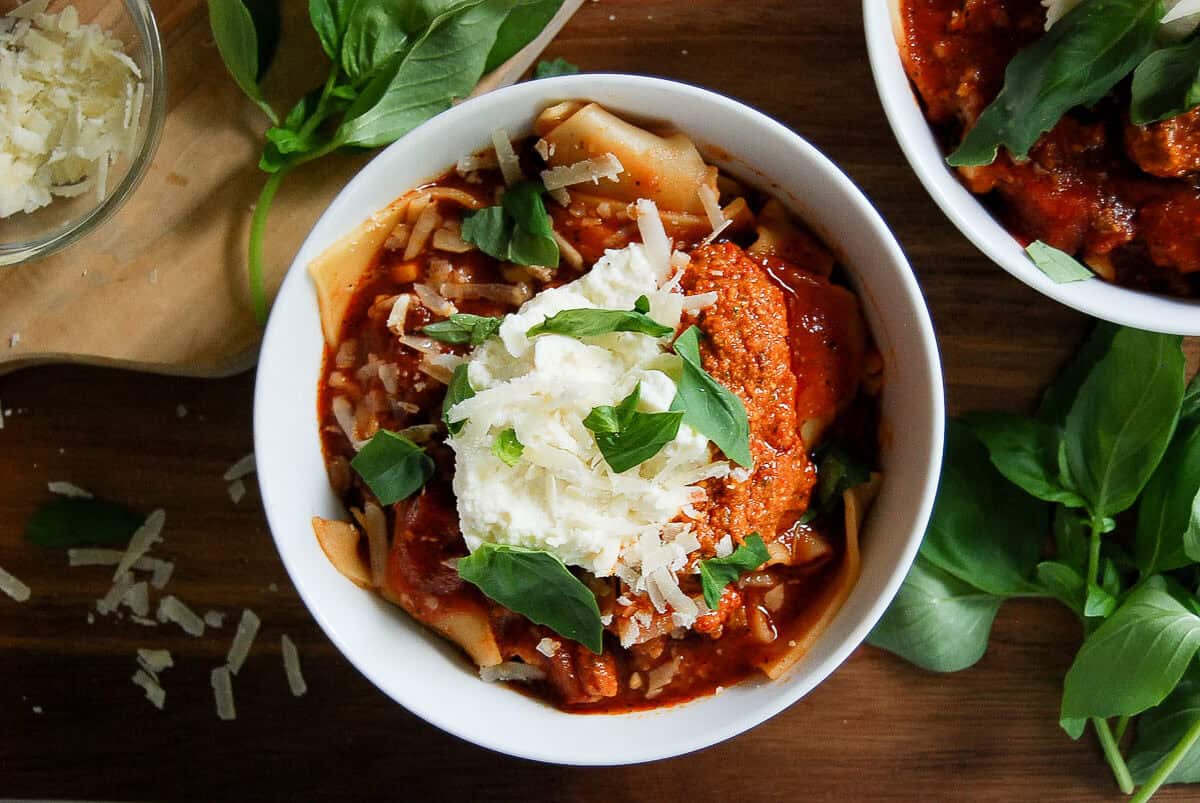 A bowl of lasagna soup topped with ricotta cheese, red pepper pesto, grated parmesan, and fresh basil leaves sits on a wooden table, surrounded by scattered cheese and basil.