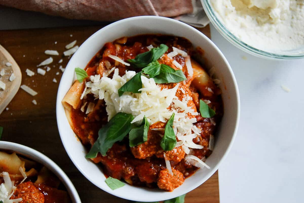 A white bowl filled with lasagna soup, topped with ricotta and red pepper pesto, grated cheese and fresh basil leaves, sits on a wooden surface next to a bowl of ricotta cheese.