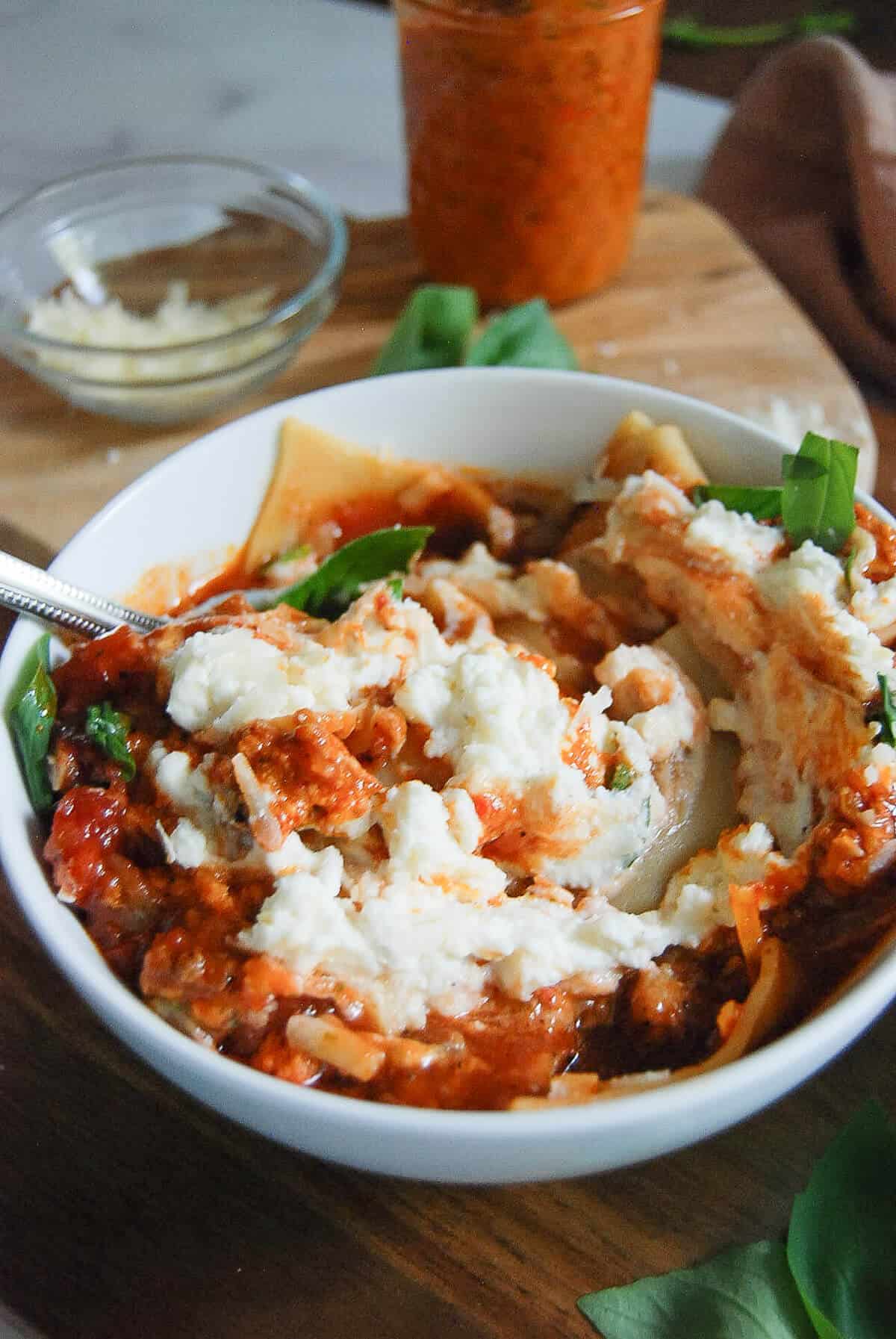 A white bowl filled with lasagna soup with ricotta cheese, red pepper pesto and fresh basil sits on a wooden surface. A fork is in the bowl, and a glass jar of sauce and a small bowl are in the background.