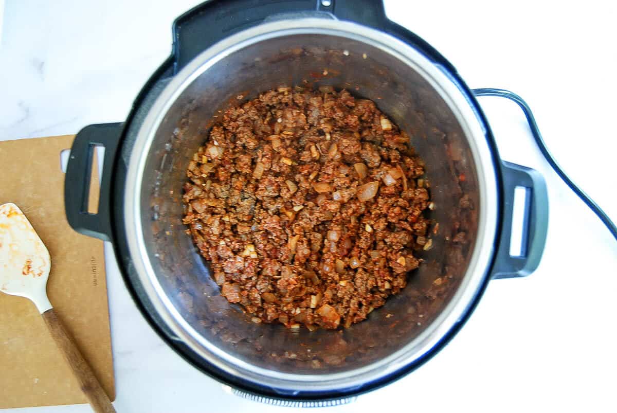 Ground meat with chopped onions and tomato sauce cooking in a slow cooker. A spatula with food residue rests on a cutting board nearby, and everything is on a white countertop.