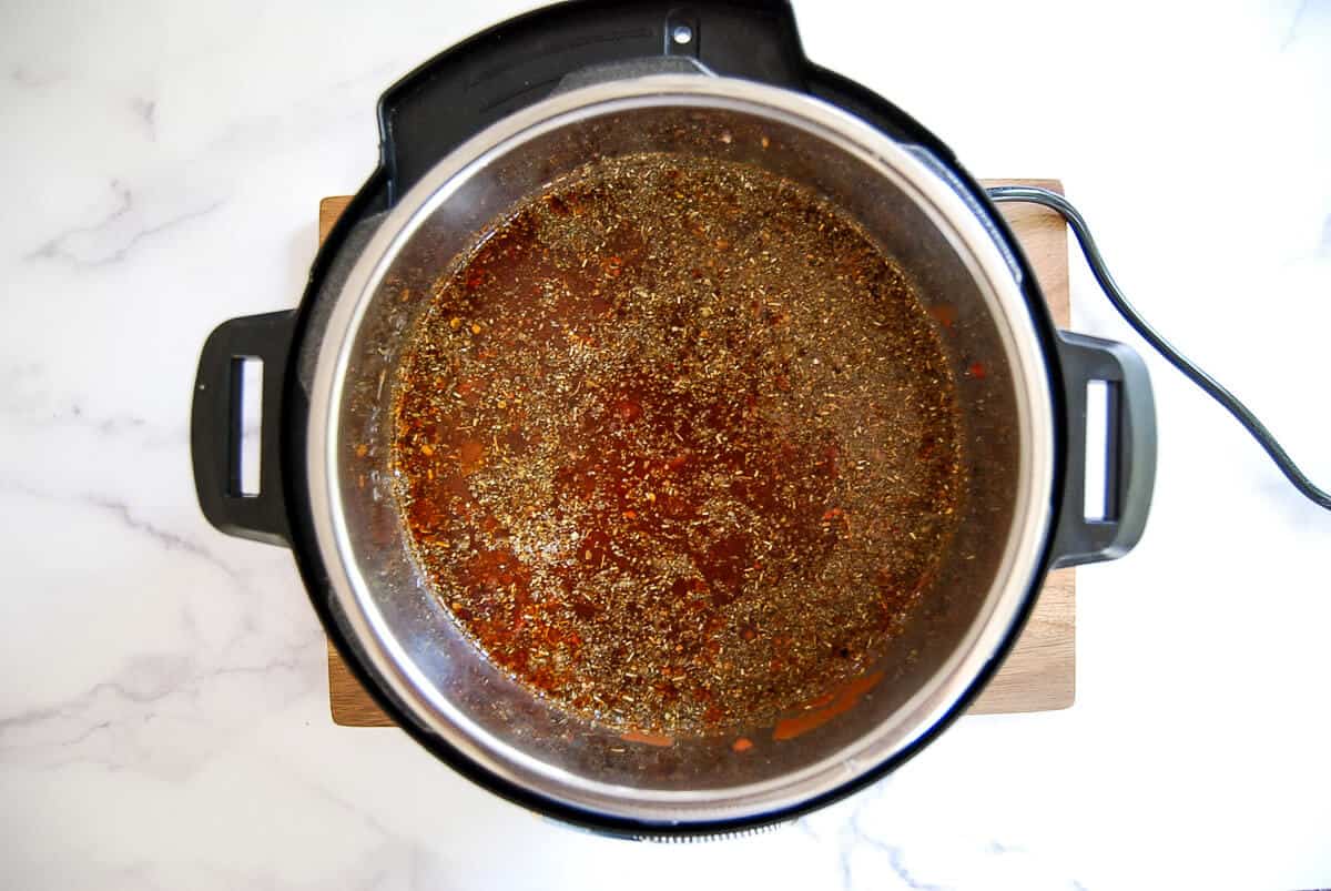 An overhead view of a crockpot, filled with seasoned soup, showing visible herbs and spices on the surface, sitting on a wooden board against a white marble background.