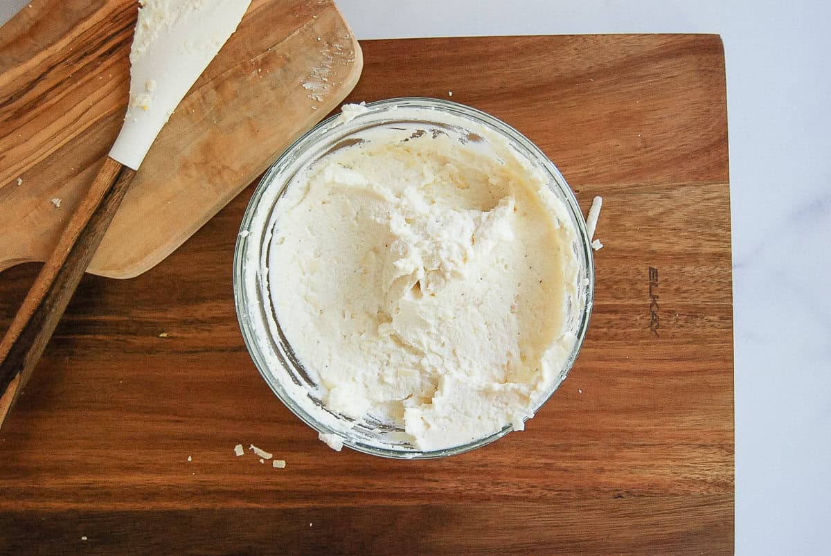 A glass bowl filled with creamy, whipped ricotta sits on a wooden cutting board, next to a spatula with some cheese on it.