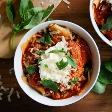 A bowl of lasagna soup, with tomato sauce, shredded cheese, a dollop of ricotta, and fresh basil leaves, with extra basil and cheese scattered nearby on a wooden surface.