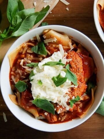 A bowl of lasagna soup, with tomato sauce, shredded cheese, a dollop of ricotta, and fresh basil leaves, with extra basil and cheese scattered nearby on a wooden surface.