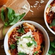 Two white bowls filled with lasagna soup, topped with ricotta cheese, red pepper pesto, and basil leaves on a wooden surface, with shredded cheese and fresh basil scattered nearby.