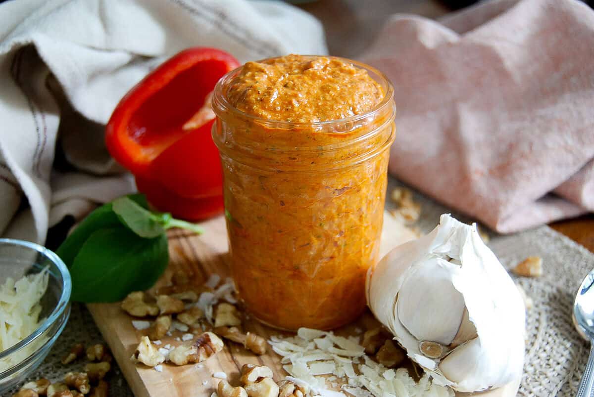 A glass jar filled with red pesto sauce sits on a wooden board, surrounded by a garlic bulb, basil leaves, grated cheese, walnuts, and half a red bell pepper. A beige cloth and bowl are in the background.
