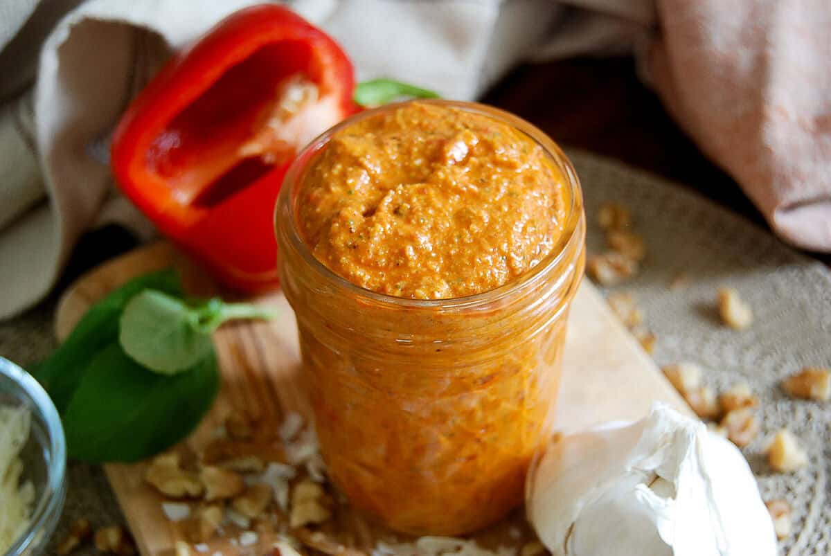 A jar of red pepper spread sits on a wooden board with fresh basil, walnuts, a halved red bell pepper, and a bulb of garlic nearby.