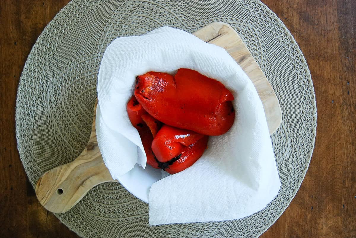 A white bowl lined with paper towel holds roasted red bell peppers on a wooden cutting board, placed on a woven placemat atop a wooden table.