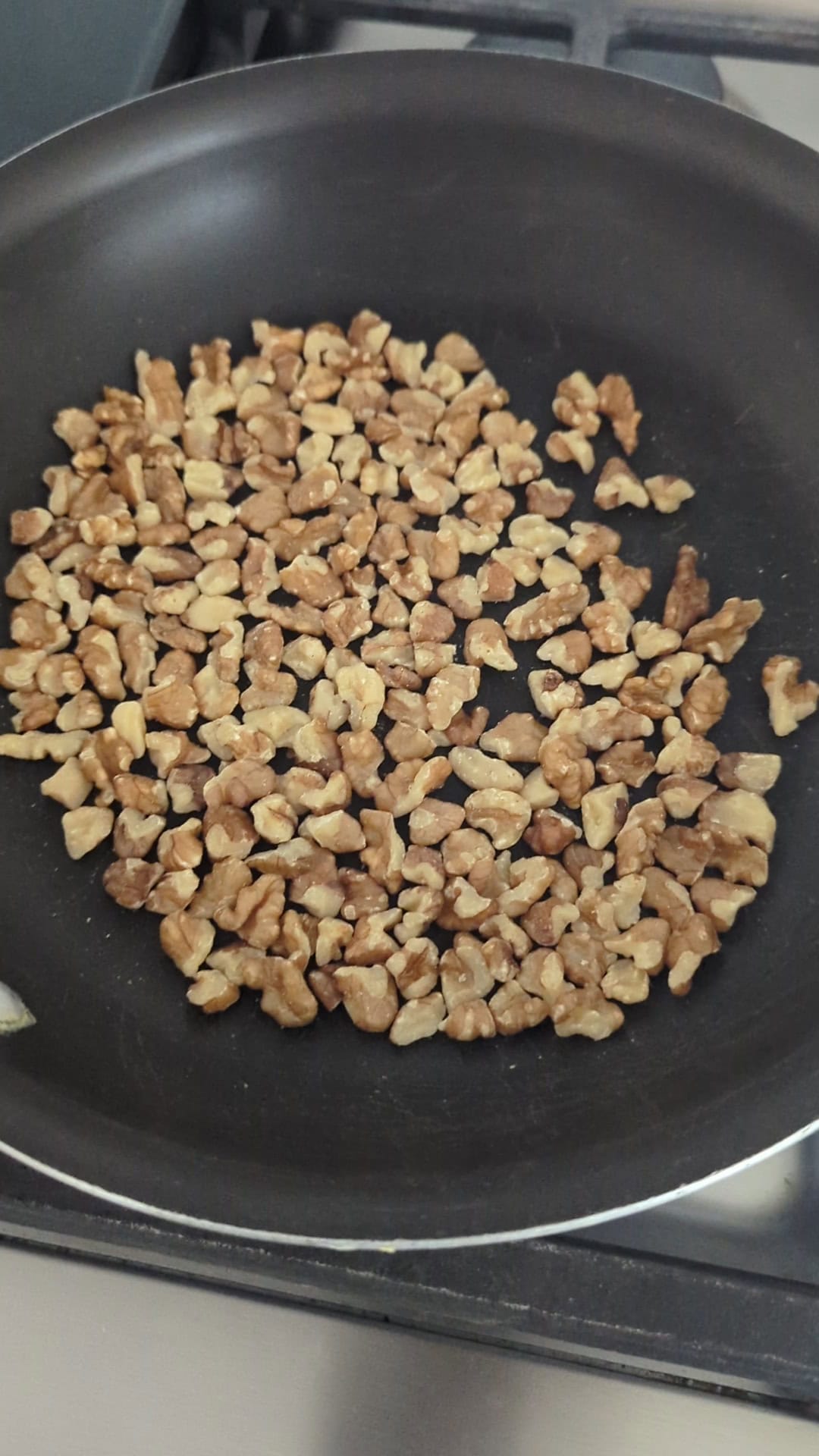 Chopped walnuts spread in a single layer on a black nonstick frying pan, being toasted on a stovetop.