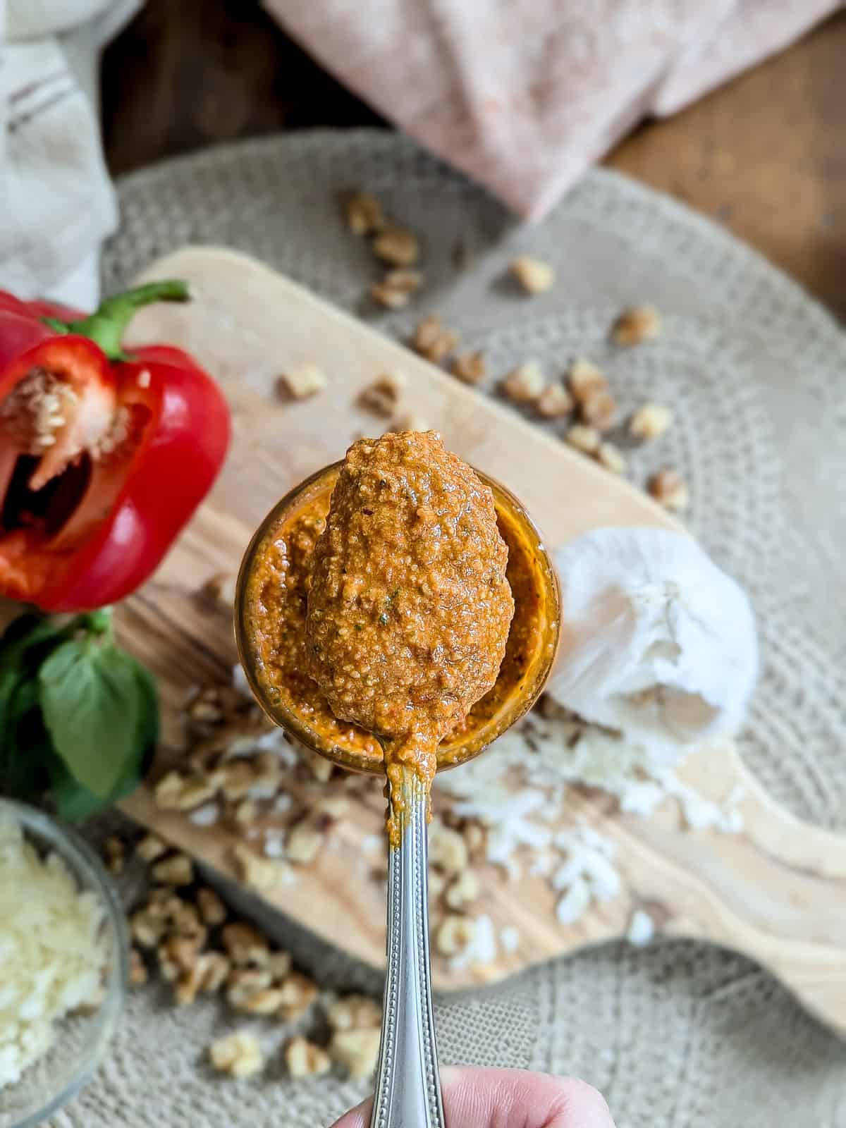 A spoonful of red pepper pesto held over a wooden board with walnuts, grated cheese, a red bell pepper, garlic, and basil in the background.