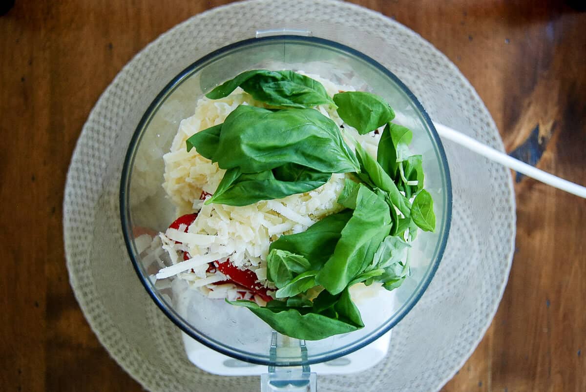 Overhead view of a food processor filled with shredded cheese, fresh basil leaves, and roasted red pepper slices on a round woven placemat.