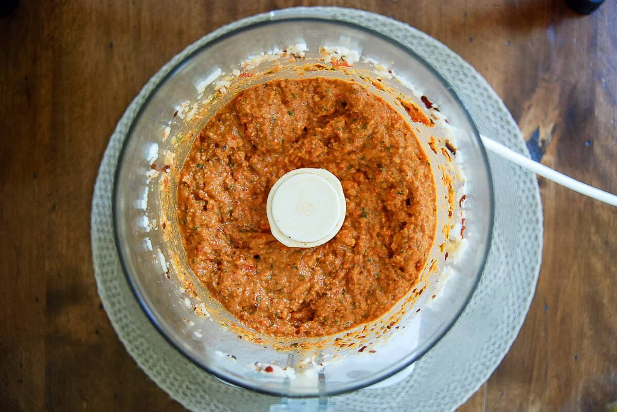 A food processor filled with a blended roasted red pepper pesto on a woven placemat on a wooden table, viewed from above.