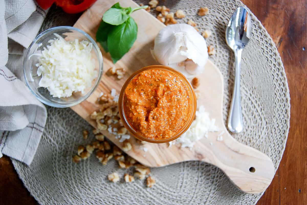 A jar of roasted red pepper pesto sauce on a wooden board, surrounded by chopped nuts, shredded cheese, a garlic bulb, basil leaves, a spoon, and a beige cloth on a woven placemat.