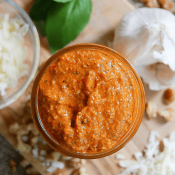 A jar of red pepper pesto sauce sits on a wooden surface, surrounded by garlic, shredded cheese, walnuts, and basil leaves.