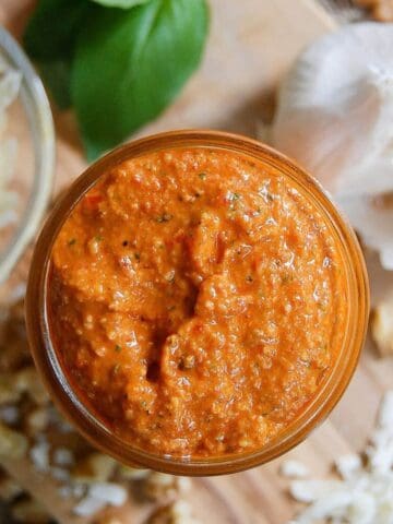 A jar of red pepper pesto sauce sits on a wooden board, surrounded by shredded cheese, walnuts, garlic, and fresh basil leaves.