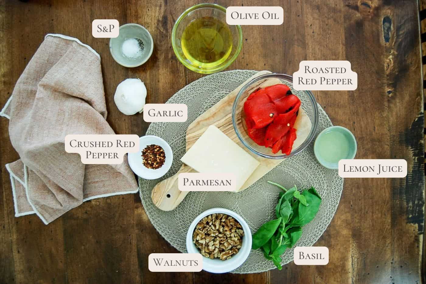 Top-down view of ingredients for roasted red pepper pesto: salt and pepper, garlic, olive oil, roasted red peppers, lemon juice, basil, walnuts, parmesan, and crushed red pepper on a wooden table.